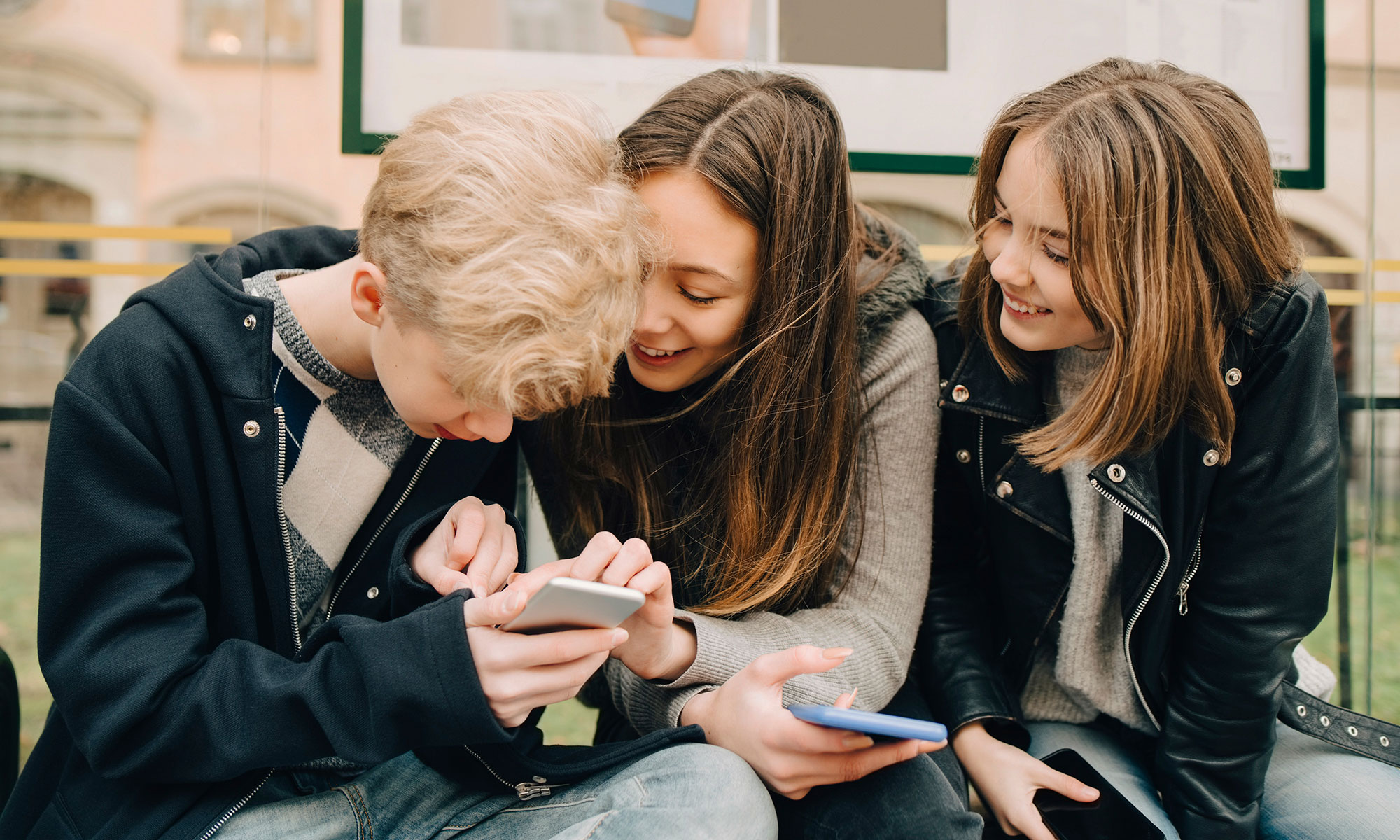 Tre ungdommar sitter på en busshållplats och tittar på sina mobiltelefoner