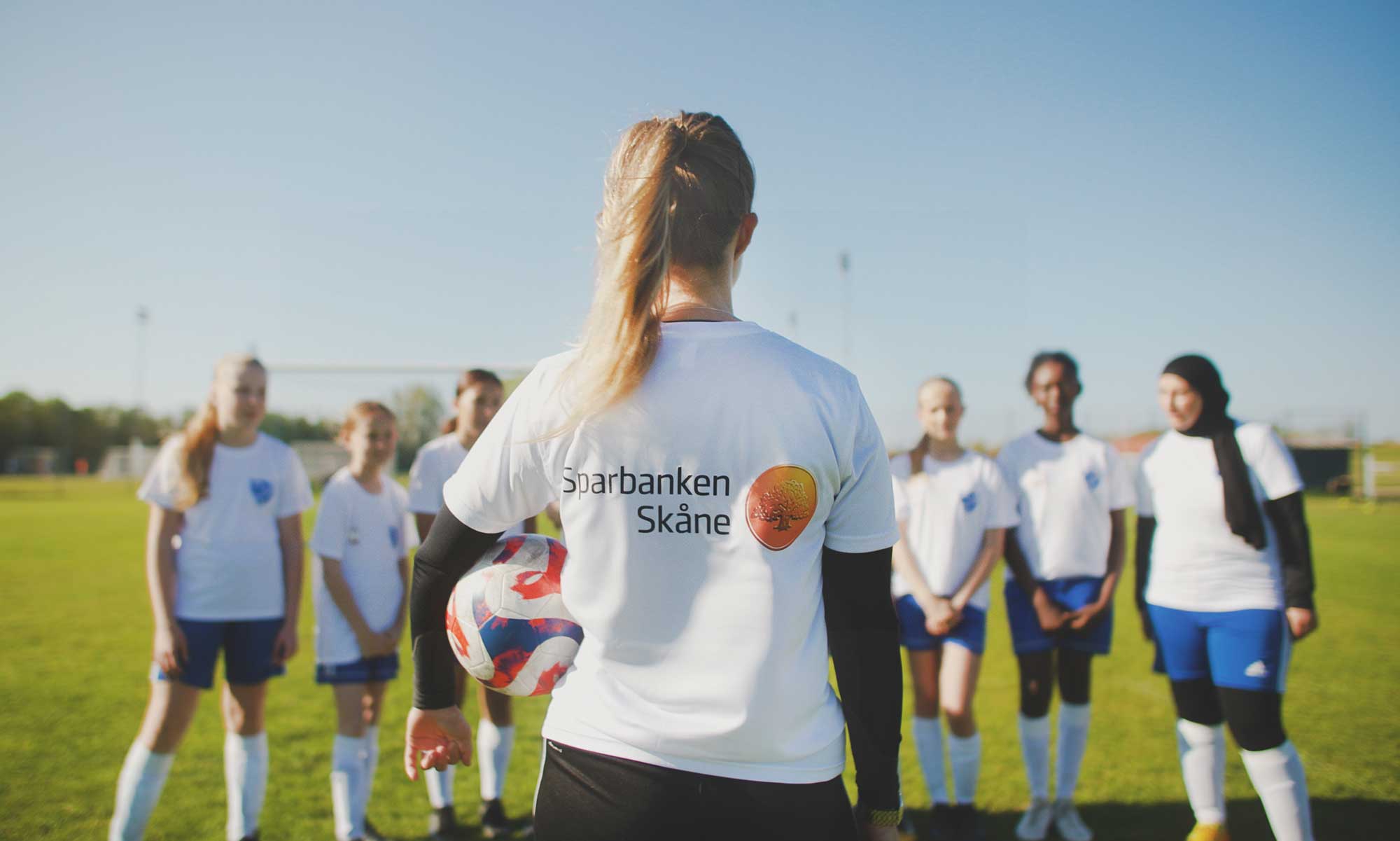 Fotbololstränare står på en fotbollsplan med ryggen vänd mot kameran och tittar på tjejer i ett fotbollslag. 